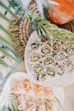 A visually appealing arrangement of sushi rolls and nigiri neatly presented on white plates. The sushi rolls contain rice, cucumber, and avocado wrapped in seaweed, while the nigiri showcases salmon and white fish topped with a drizzle of sauce. The composition includes decorative green leaves and a woven placemat adding a natural, fresh touch.