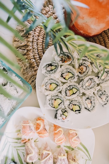 A visually appealing arrangement of sushi rolls and nigiri neatly presented on white plates. The sushi rolls contain rice, cucumber, and avocado wrapped in seaweed, while the nigiri showcases salmon and white fish topped with a drizzle of sauce. The composition includes decorative green leaves and a woven placemat adding a natural, fresh touch.