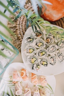A visually appealing arrangement of sushi rolls and nigiri neatly presented on white plates. The sushi rolls contain rice, cucumber, and avocado wrapped in seaweed, while the nigiri showcases salmon and white fish topped with a drizzle of sauce. The composition includes decorative green leaves and a woven placemat adding a natural, fresh touch.