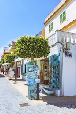 A charming street scene with bright and colorful buildings and shops. One can see a tree with a leafy canopy, various signs with beach-themed messages, and windows with blue shutters. The street is lined with small businesses displaying merchandise such as clothing and accessories. A clear blue sky complements the vibrant atmosphere.