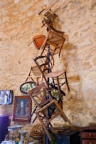 A collection of wooden chairs is stacked vertically against a rustic stone wall. The arrangement is artistically balanced, with chairs appearing to be attached to a central pole. The background includes framed artwork and decorative items, contributing to an eclectic and vintage atmosphere.