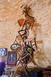 A collection of wooden chairs is stacked vertically against a rustic stone wall. The arrangement is artistically balanced, with chairs appearing to be attached to a central pole. The background includes framed artwork and decorative items, contributing to an eclectic and vintage atmosphere.