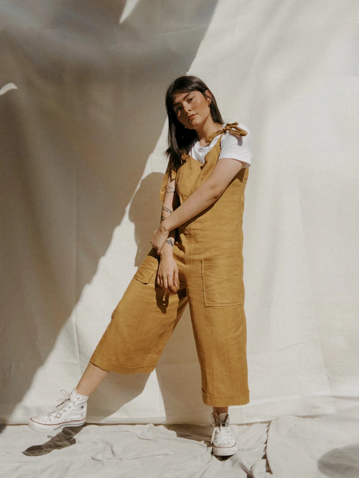 Woman in mustard canvas overalls and a white tee against a draped backdrop