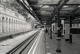 A black and white photograph of a quiet train station, featuring multiple tracks leading into a tunnel in the distance. The platform is partially covered by an architectural structure with columns and a roof. A person is standing on the platform, looking at something in their hands, possibly a phone, while wearing a backpack. The left side of the image shows brick arches and foliage, adding a historic feel.
