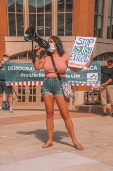 A woman holds a megaphone and a sign reading 'Stop Abortion Extremism' at a public demonstration. Other participants in the background hold a banner that reads 'Democrats for Life of America' with additional people standing nearby. The setting appears to be an urban area with architectural elements.