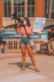 A woman holds a megaphone and a sign reading 'Stop Abortion Extremism' at a public demonstration. Other participants in the background hold a banner that reads 'Democrats for Life of America' with additional people standing nearby. The setting appears to be an urban area with architectural elements.