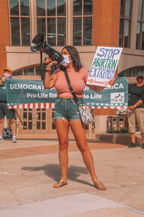 A woman holds a megaphone and a sign reading 'Stop Abortion Extremism' at a public demonstration. Other participants in the background hold a banner that reads 'Democrats for Life of America' with additional people standing nearby. The setting appears to be an urban area with architectural elements.