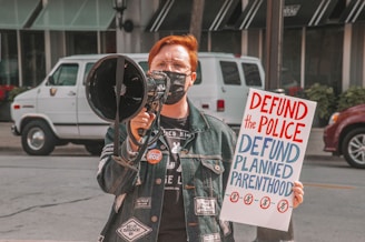 A person wearing a denim jacket and a face mask is holding a megaphone and a protest sign. The sign has bold red and blue letters reading 'DEFUND the POLICE' and 'DEFUND PLANNED PARENTHOOD'. In the background, there are vehicles parked on the street and a building with large windows.