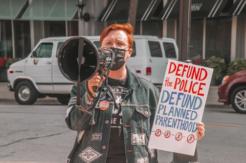 A person wearing a denim jacket and a face mask is holding a megaphone and a protest sign. The sign has bold red and blue letters reading 'DEFUND the POLICE' and 'DEFUND PLANNED PARENTHOOD'. In the background, there are vehicles parked on the street and a building with large windows.