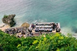 An outdoor bar is situated on a cliff overlooking the ocean, with people sitting around tables enjoying the view. The area is surrounded by lush greenery and large rocks partially submerged in the water.