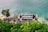 An outdoor bar is situated on a cliff overlooking the ocean, with people sitting around tables enjoying the view. The area is surrounded by lush greenery and large rocks partially submerged in the water.