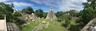 A panoramic view of an ancient archaeological site featuring a large stepped pyramid surrounded by lush greenery and smaller stone structures. The sky above is partly cloudy, casting soft shadows on the ground.
