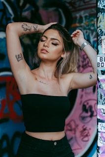 A person with tattoos on their arms and chest poses with arms raised and eyes closed, set against a vibrant graffiti-covered wall. The individual is wearing a black tube top and has a nose ring and wristwatch.