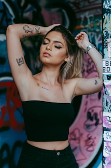 A person with tattoos on their arms and chest poses with arms raised and eyes closed, set against a vibrant graffiti-covered wall. The individual is wearing a black tube top and has a nose ring and wristwatch.