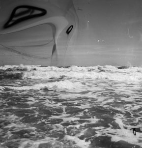 A black and white image depicting a turbulent ocean with frothy waves under a clear sky. The surface of the water is dynamic, with waves crashing and spreading foam. There are abstract, #B8C0CC black shapes overlaying the image, giving it a surreal and dreamlike quality.