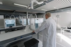 A person wearing a lab coat is inspecting a 3D printer in a modern, well-lit laboratory setting. The room has multiple 3D printers placed on shelves, with visible ventilation ducts and a clean, organized environment. The light from the overhead fixtures and windows highlights the mechanical equipment.