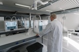 A person wearing a lab coat is inspecting a 3D printer in a modern, well-lit laboratory setting. The room has multiple 3D printers placed on shelves, with visible ventilation ducts and a clean, organized environment. The light from the overhead fixtures and windows highlights the mechanical equipment.
