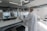 A person wearing a lab coat is inspecting a 3D printer in a modern, well-lit laboratory setting. The room has multiple 3D printers placed on shelves, with visible ventilation ducts and a clean, organized environment. The light from the overhead fixtures and windows highlights the mechanical equipment.