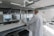 A person wearing a lab coat is inspecting a 3D printer in a modern, well-lit laboratory setting. The room has multiple 3D printers placed on shelves, with visible ventilation ducts and a clean, organized environment. The light from the overhead fixtures and windows highlights the mechanical equipment.