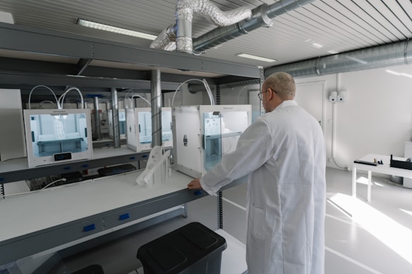 A person wearing a lab coat is inspecting a 3D printer in a modern, well-lit laboratory setting. The room has multiple 3D printers placed on shelves, with visible ventilation ducts and a clean, organized environment. The light from the overhead fixtures and windows highlights the mechanical equipment.