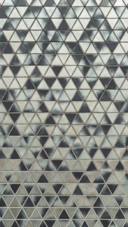 A geometric pattern featuring numerous small triangles arranged in a repetitive, tessellated format. The surface appears textured, with varying shades of grey and black giving a gradient or shadow effect across the image.