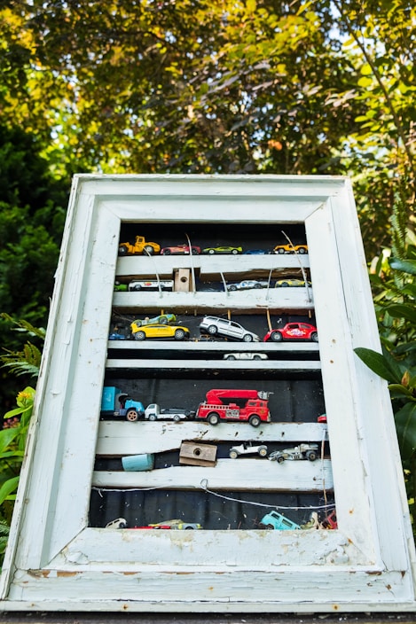 A collection of miniature toy cars and vehicles arranged on shelves inside a weathered, white-framed shadow box. The display is outdoors, surrounded by green foliage and trees, with sunlight filtering through the leaves.