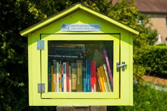 A small, yellow wooden box functions as a free library, filled with various books visible through a glass door. It is structured like a miniature house with a slanted roof and a sign that reads 'Little Free Library'. Positioned outdoors, it's surrounded by lush greenery.