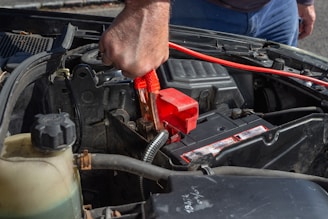 A person is using a red jumper cable to connect to a car battery. The engine compartment is visible, with various car parts surrounding the battery. The person’s hand is gripping the cable tightly, suggesting they are in the process of jump-starting the vehicle.