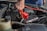 A person is using a red jumper cable to connect to a car battery. The engine compartment is visible, with various car parts surrounding the battery. The person’s hand is gripping the cable tightly, suggesting they are in the process of jump-starting the vehicle.