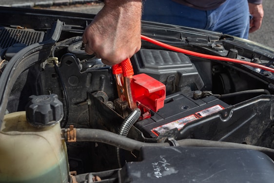 A person is using a red jumper cable to connect to a car battery. The engine compartment is visible, with various car parts surrounding the battery. The person’s hand is gripping the cable tightly, suggesting they are in the process of jump-starting the vehicle.