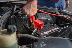 A person is using a red jumper cable to connect to a car battery. The engine compartment is visible, with various car parts surrounding the battery. The person’s hand is gripping the cable tightly, suggesting they are in the process of jump-starting the vehicle.