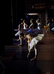 A group of ballerinas is preparing backstage, dressed in elegant tutus with intricate gold detailing. They stand in a dimly lit area with shadows and muted lighting creating a serene atmosphere. Each dancer appears focused and graceful, ready to perform.
