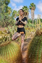 A person with tattoos practices yoga in a desert garden setting surrounded by various types of cacti and palm trees, under a bright blue sky.