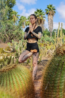 A person with tattoos practices yoga in a desert garden setting surrounded by various types of cacti and palm trees, under a bright blue sky.