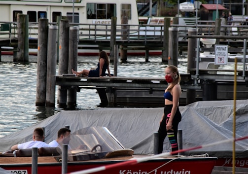 At a marina, two men are seated in a motorboat while a woman stands nearby wearing colorful leggings and a red face mask. In the background, another woman is sitting on a wooden dock, and there are several boats and piers visible.