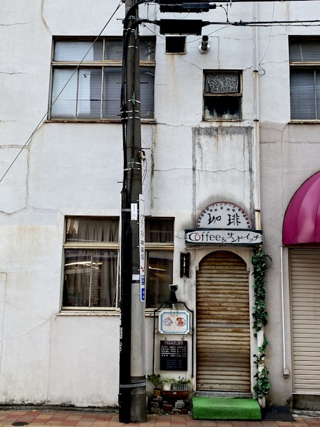 A narrow white building exterior with a closed wooden shutter door, marked by a sign above that reads 'Coffee & Sandwich.' The facade shows signs of aging with visible cracks and discoloration. A vertical black utility pole stands in front, adorned with various wires and a small sign, along with potted plants at the base of the building.