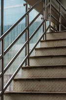 An upward view of a metal staircase with diamond plate treads and stainless steel railings. The staircase is next to a large glass window with raindrops on it, suggesting wet or rainy weather outside.