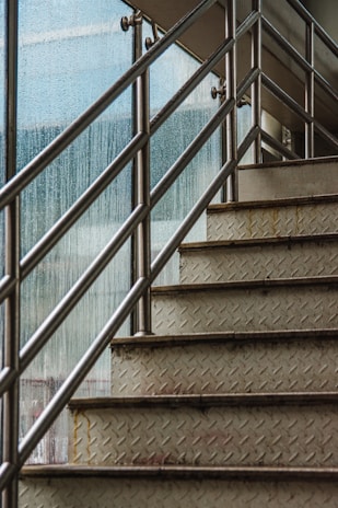 An upward view of a metal staircase with diamond plate treads and stainless steel railings. The staircase is next to a large glass window with raindrops on it, suggesting wet or rainy weather outside.