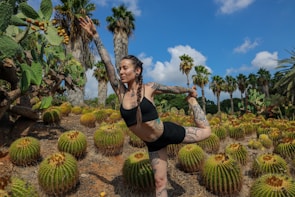 A person with tattoos is practicing a yoga pose in a desert-like setting surrounded by cacti and palm trees. The sky is clear with some clouds, adding to the vibrant and serene atmosphere.
