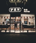 A pastry shop is captured at night with its illuminated signage. Inside, a few people are seen standing and interacting at the counter, likely ordering or purchasing pastries. The interior is well-lit, showing a clean and modern design with wooden accents and a variety of pastries on display.