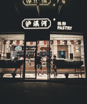 A pastry shop is captured at night with its illuminated signage. Inside, a few people are seen standing and interacting at the counter, likely ordering or purchasing pastries. The interior is well-lit, showing a clean and modern design with wooden accents and a variety of pastries on display.