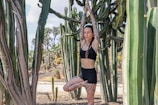A woman with tattoos is performing a yoga pose, standing on one leg with her arms raised above her head. She is surrounded by tall cacti in a desert-like setting. The woman is wearing a black sports bra and shorts.