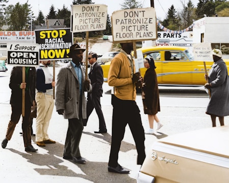 A group of people are marching on a street, holding signs advocating against racial discrimination and supporting open housing. The signs display messages such as 'Stop Racial Discrimination Now' and 'I Support Open Housing'. The marchers are diverse in terms of gender and race, and the scene is set in a suburban area with houses and a workshop visible in the background. A bright yellow car is passing by in the background.