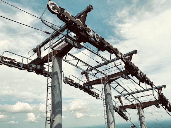 A partial view of a ski lift mechanism with large wheels and cables, set against a cloudy sky. The metallic structure includes ladders, platforms, and pulleys, showcasing an intricate engineering design.