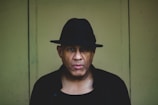 A person is wearing a black hat and dark clothing, standing against a plain, muted green background. The lighting is low, creating a shadowy and mysterious ambiance.