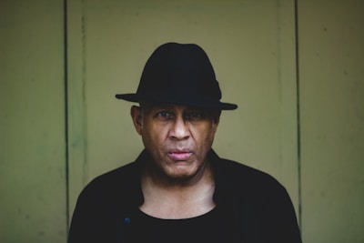 A person is wearing a black hat and dark clothing, standing against a plain, muted green background. The lighting is low, creating a shadowy and mysterious ambiance.