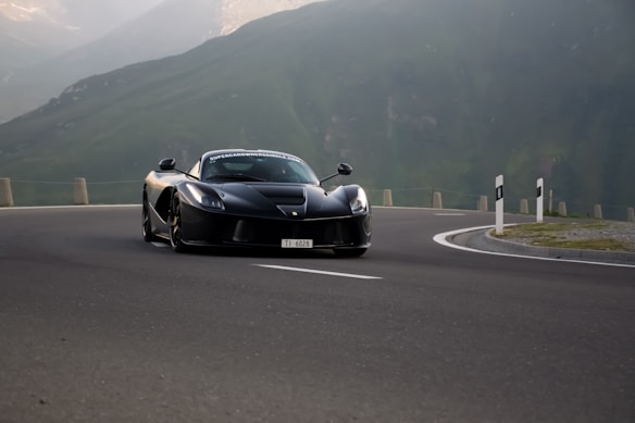A sleek black Ferrari sports car is navigating a mountain road with grassy hills and a cloudy sky in the background. The vehicle is positioned slightly towards the camera, emphasizing its aerodynamic design and shiny surface as it speeds down the curvy highway.