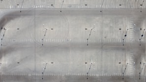 An aerial view of a large, open concrete or asphalt surface. The surface is marked with various lines and circles, possibly indicating parking spaces or runway guidance. Light poles with small shadows are evenly distributed across the area.