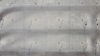 An aerial view of a large, open concrete or asphalt surface. The surface is marked with various lines and circles, possibly indicating parking spaces or runway guidance. Light poles with small shadows are evenly distributed across the area.