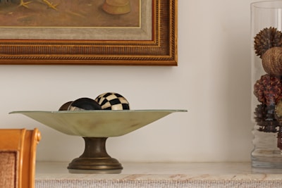 A decorative setup featuring a pedestal bowl with black and white checkered and solid black spheres inside it. To the right, a transparent glass container holds natural elements like pine cones. The background includes a framed painting with a golden brown frame.